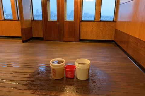 Two white buckets and a red pail catching drips on a wet wooden floor in a wood paneled room