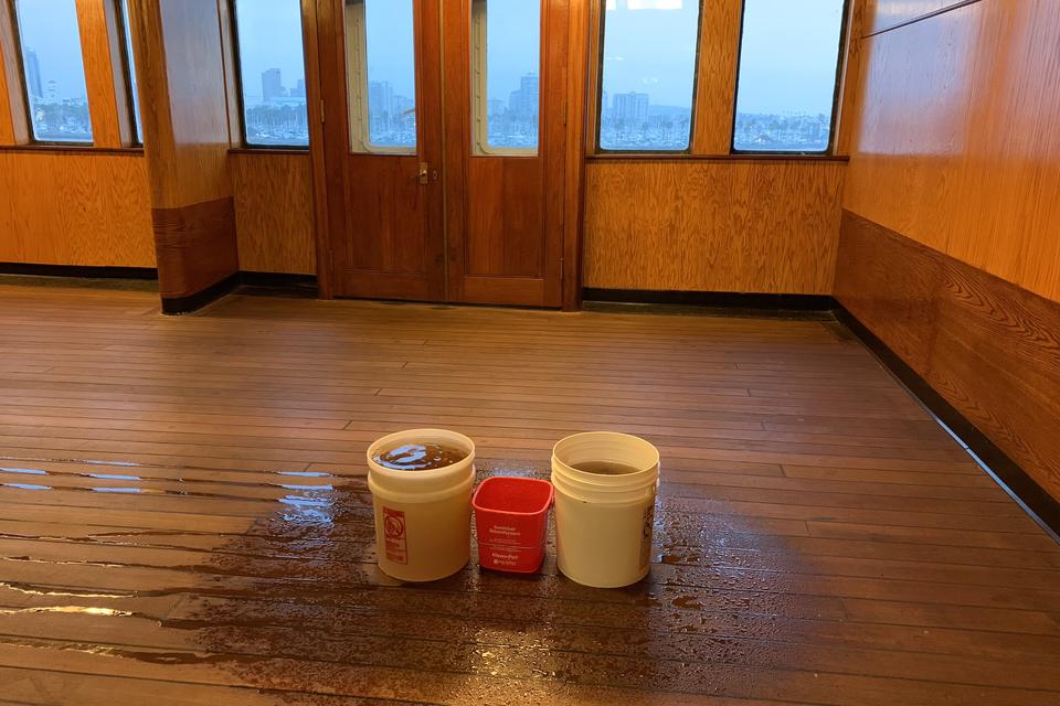 Two white buckets and a red pail catching drips on a wet wooden floor in a wood paneled room