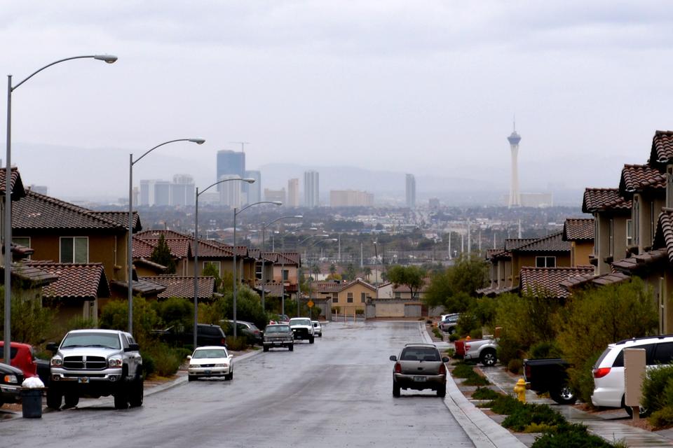 A wet residential street lined with tile roof houses and parked trucks, the Strip and Stratosphere tower in haze beyond