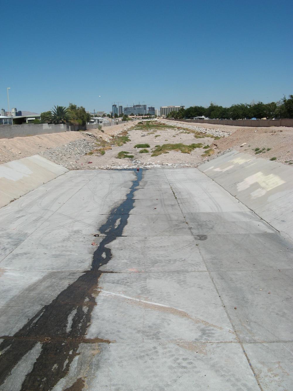 A dry concrete flood channel with a thin dark trickle down the middle and high rise towers on the horizon