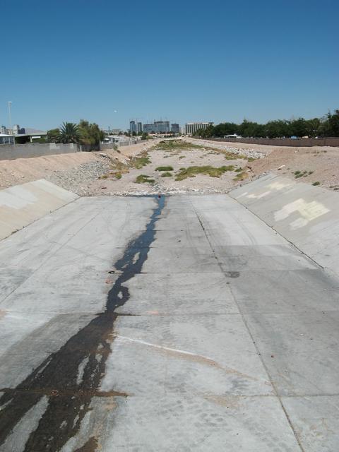 A dry concrete flood channel with a thin dark trickle down the middle and high rise towers on the horizon