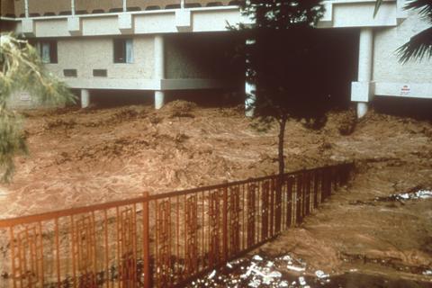 Muddy floodwater rushing past an orange railing and under a raised hotel building
