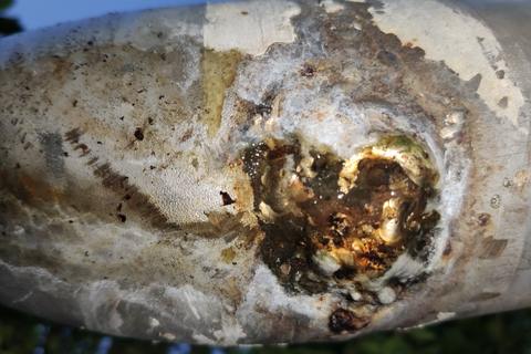 Close view from below of a corroded hole in a pipe bend, wet with rust and mineral crust